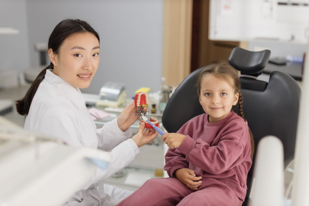 Happy female dentist tells little child girl how to brush teeth on artificial jaw model