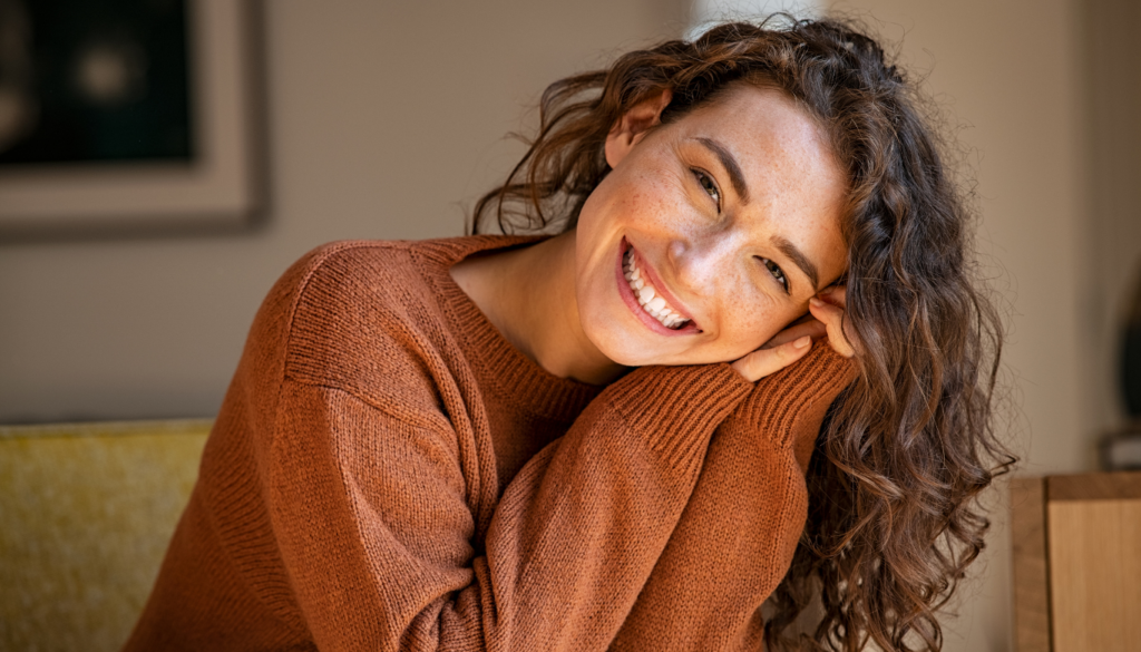 Smiling woman with long wavy hair leaning on her hands, wearing a rust-colored knit sweater indoors.