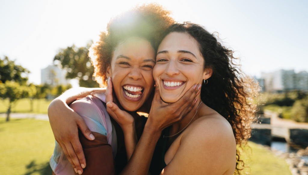Two women of different backgrounds hug and laugh together in a sunny park, faces close up.