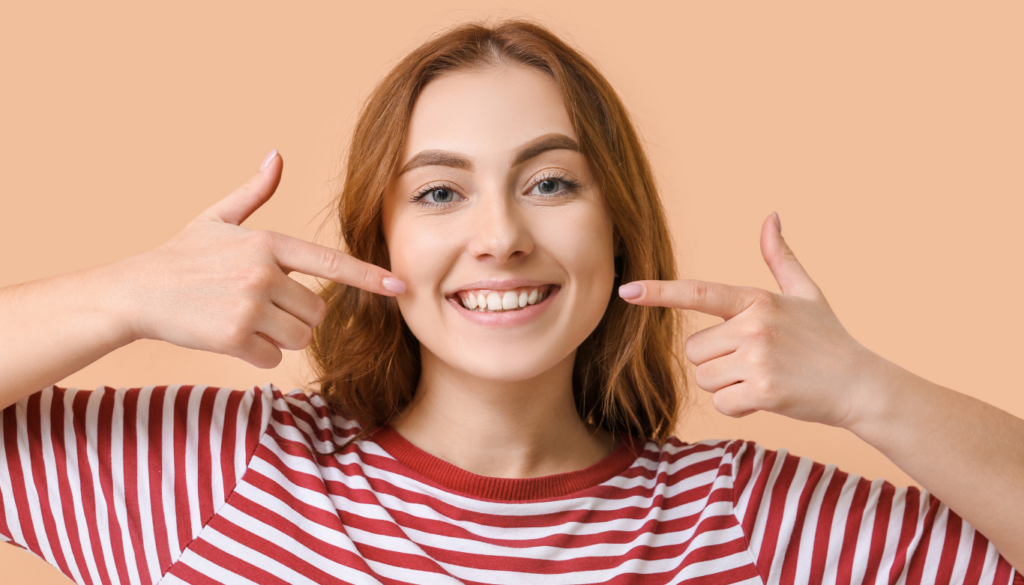 dana-street-dental-wisdom-teeth | Dana Street Dental Smiling woman with red hair in a red-and-white striped shirt points to her teeth with both hands against a peach background.