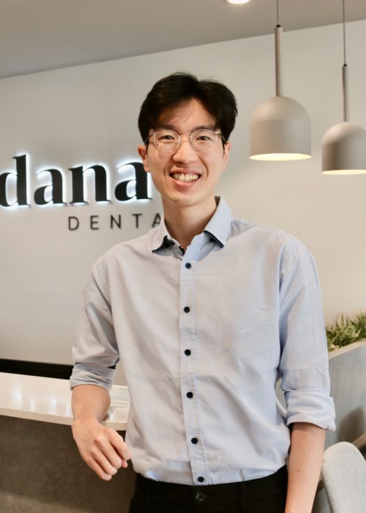 Smiling man in a light blue button-down shirt standing at a dental clinic reception with a sign behind him reading 'DENTAL'
