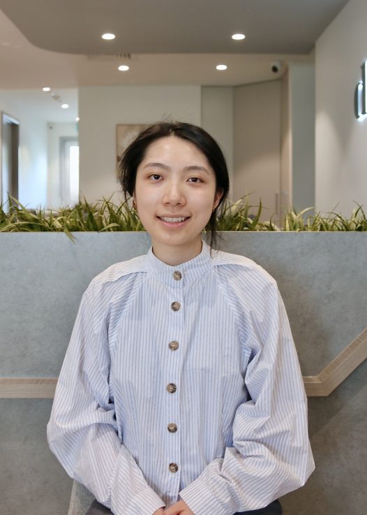 Smiling woman in a light blue striped blouse seated in a modern indoor space with a green planter behind her.