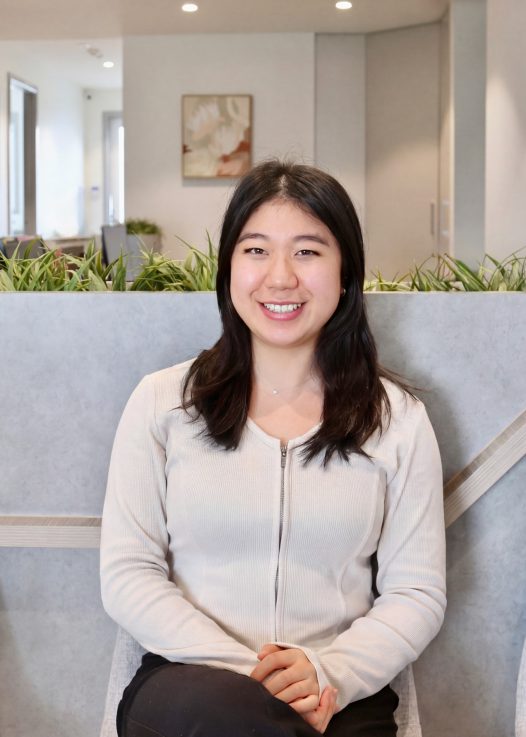 Smiling woman seated indoors with hands folded in her lap, beige cardigan, office backdrop and green plants behind her.